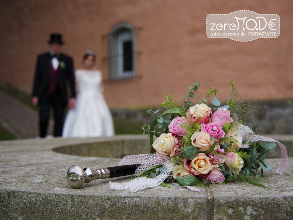 Brautstrauss aus rosa und gelben Rosen mit Spazierstock des Bräutigams, im Hintergrund das Hochzeitspaar