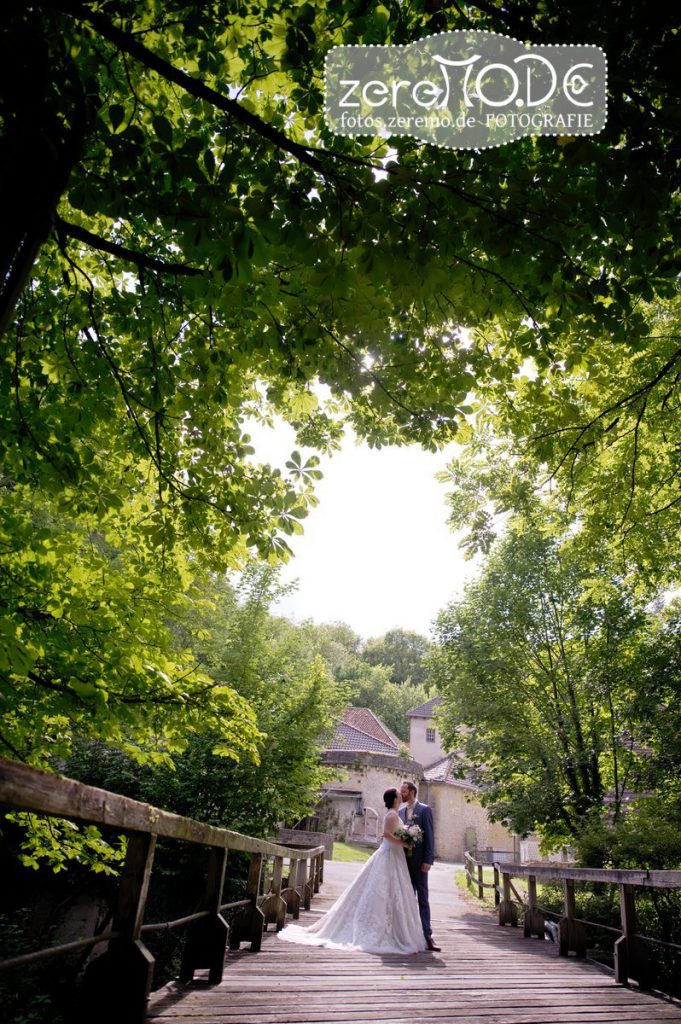 Hochzeitspaar küsst sich in der Ferne auf einer Holzbrücke unter Kastanien